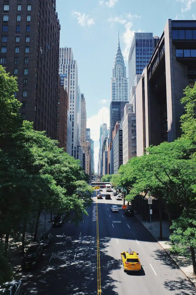 Yellow NYC taxi driving through Manhattan under green trees with the Chrysler building in the background